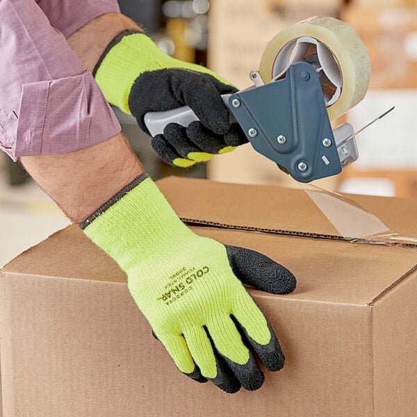 A hand wearing a Cordova Cold Snap Hi-Vis green glove with black latex palm coating using a tape dispenser to seal a box.