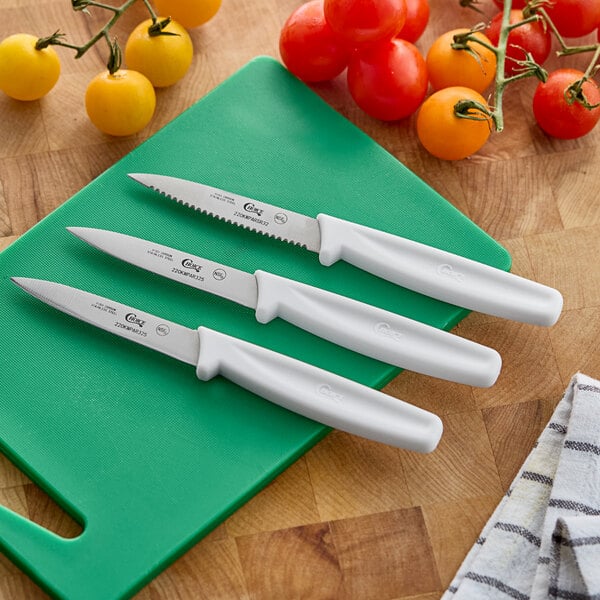 A set of three paring knives with white handles, including one serrated and two smooth edge blades, displayed on a green cutting board.