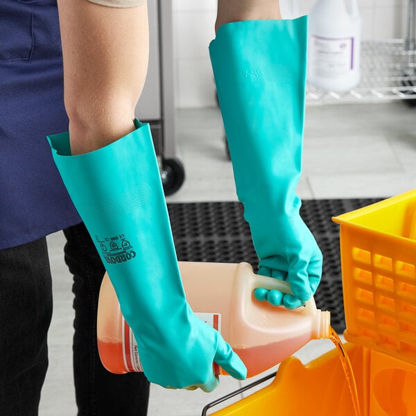 A person wearing green Cordova dishwashing gloves pouring liquid into a container.