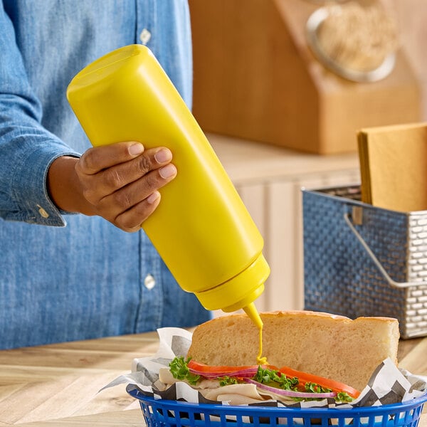 A large yellow squeeze bottle being used to dispense condiment onto a sandwich.