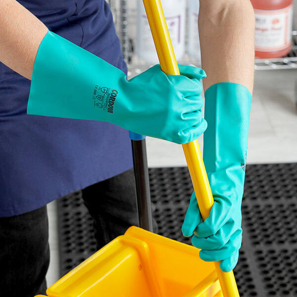 A person wearing green Cordova nitrile gloves holding a mop and a yellow bucket.