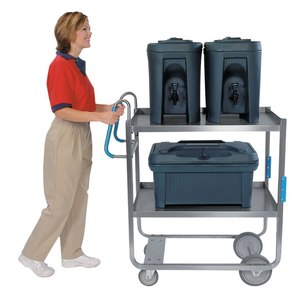 A woman in a red shirt and khaki pants pushing a Lakeside stainless steel utility cart with two containers.