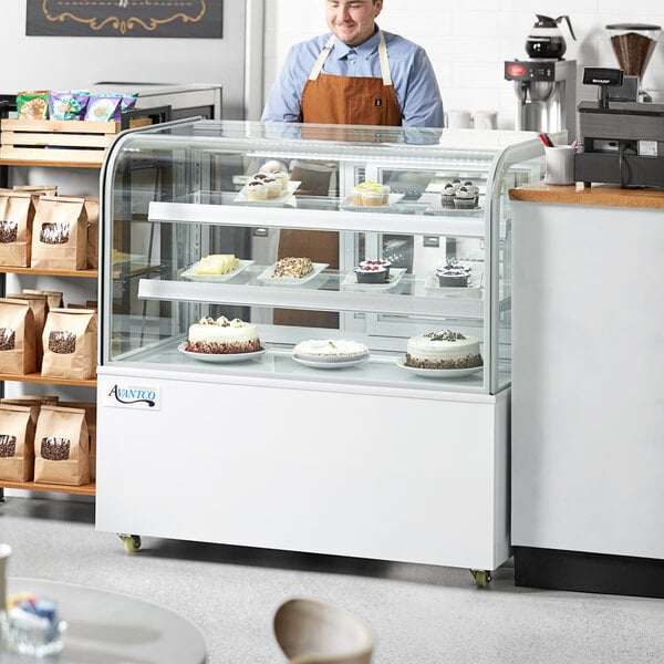 A man standing behind a white Avantco refrigerated bakery display case with cakes.