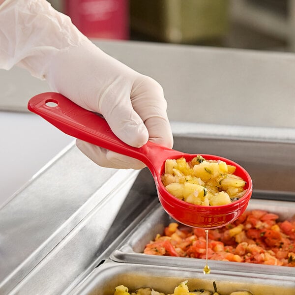 A gloved hand using a Carlisle red plastic short handle perforated portion spoon to serve food.