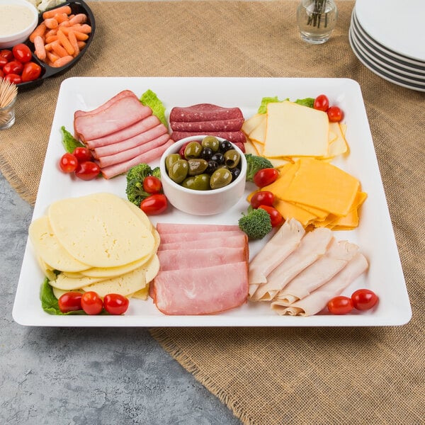 A white Fineline square plastic catering tray with meat, cheese, olives and tomatoes on a table.