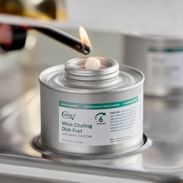 A metal canister labeled 'Wick Chafing Dish Fuel' is being lit with a lighter, with a visible flame touching the wick.
