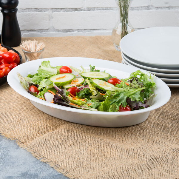 A bowl of salad in a white Fineline oval catering bowl on a table next to plates and forks.