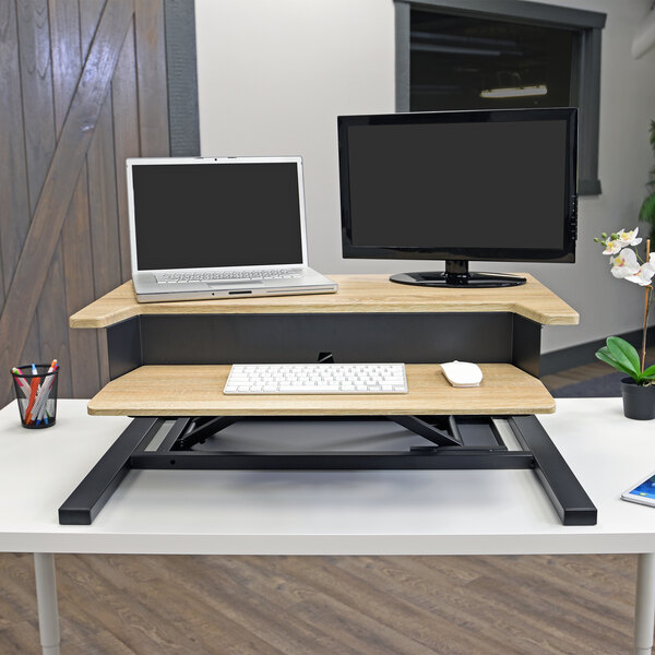 A Luxor white oak stand up desktop desk with a laptop and monitor on the top tier.