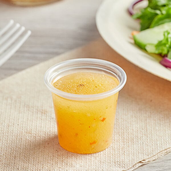 A small clear plastic portion cup filled with a yellow-orange liquid, placed on a napkin next to a plate of salad.