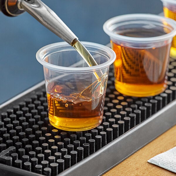 A clear plastic 1.25 oz. shot glass or portion cup filled with amber liquid, placed on a bar mat.