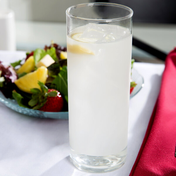 A Libbey clear frosted zombie glass filled with water and ice with a lemon slice on the rim on a table with a bowl of fruit and salad.