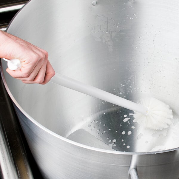 A hand holding a Carlisle white multi-purpose cleaning brush in a pot of liquid.