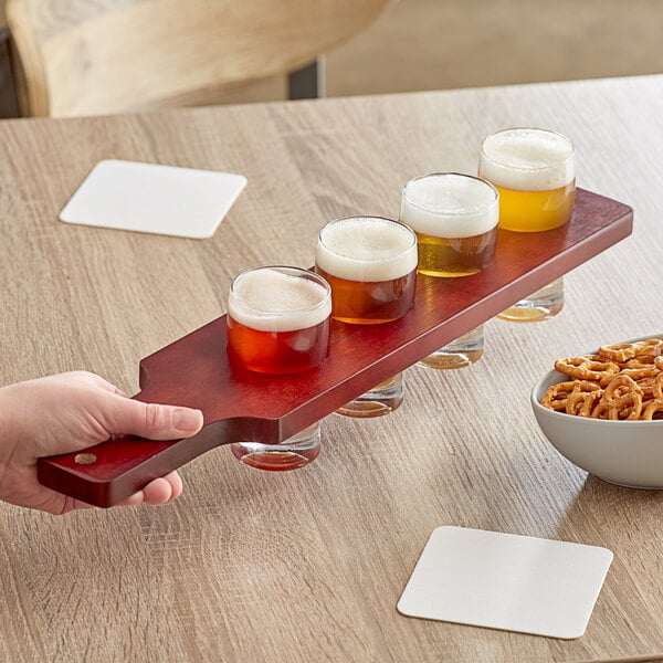 A hand holding an Acopa mahogany finish wooden flight paddle with beer glasses and a bowl of pretzels.