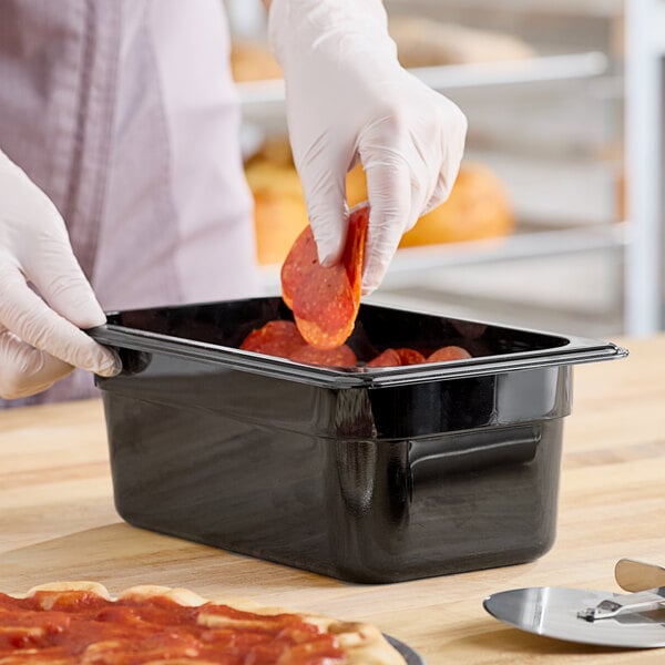 A person in gloves putting pepperoni into a black Cambro plastic food container.