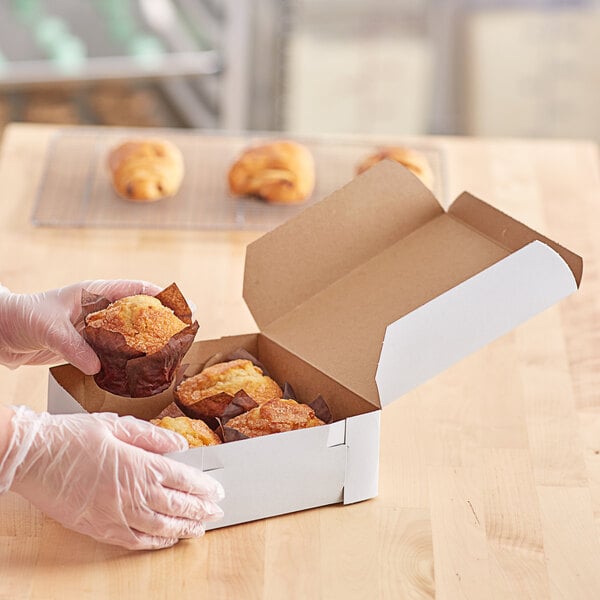 A person in gloves holding a white bakery box of muffins.