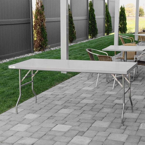 A Regency stainless steel folding work table with metal legs on a stone patio.