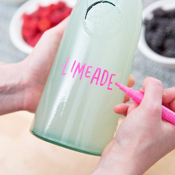 A close-up of a hand holding a Franmara neon pink mini tip glass marker and writing on a bottle.