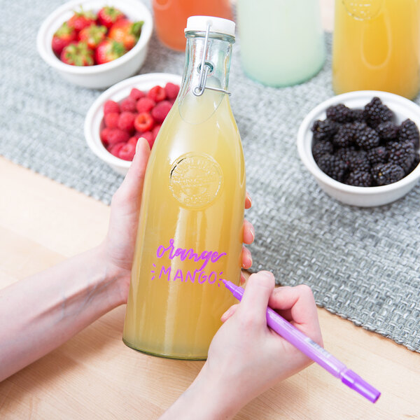 A person using a Franmara neon purple mini tip glass marker to write on a bottle of yellow liquid.