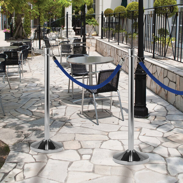 An American Metalcraft polished chrome flat head crowd control stanchion on a stone surface with a blue rope barrier.