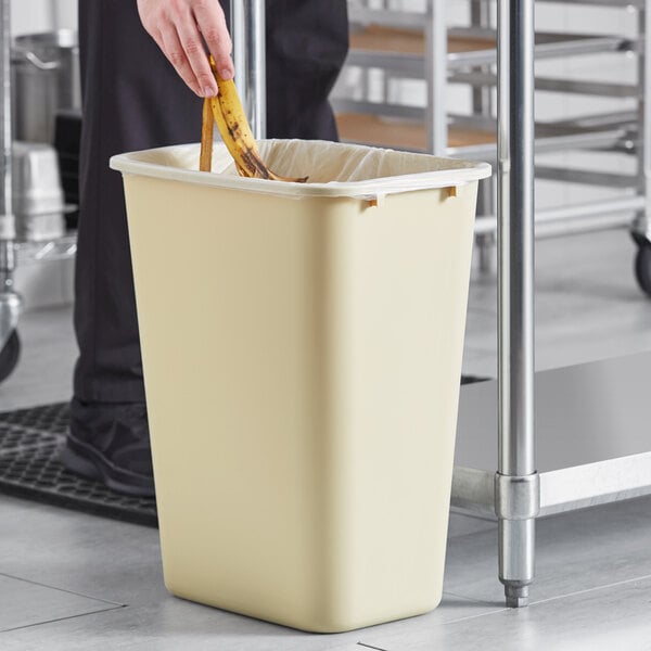 A hand putting a banana in a beige rectangular trash can.