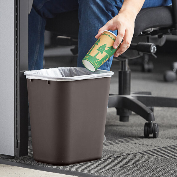 A hand holding a paper cup putting it in a Lavex brown rectangular wastebasket.