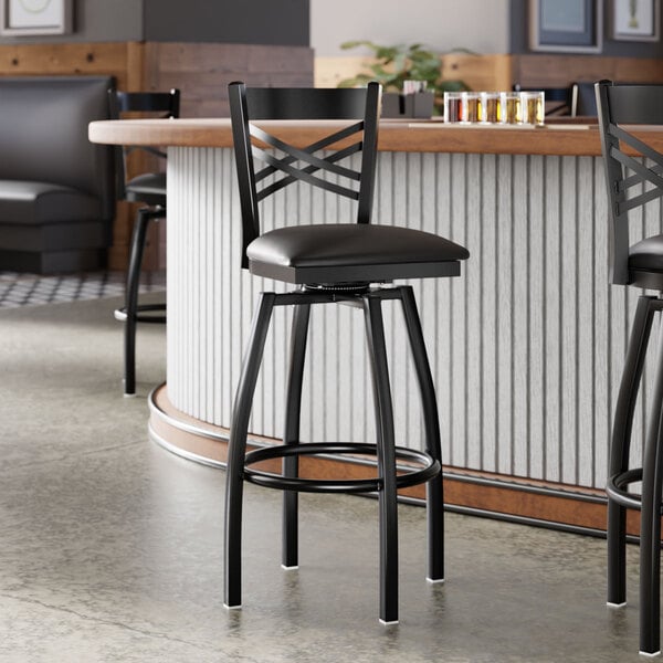 A black metal bar stool with a cross back design and a black vinyl padded swivel seat.