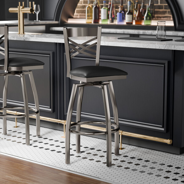 A metal bar stool with a cross back design, clear coat finish, and a black vinyl padded swivel seat.