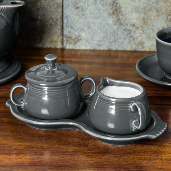 A grey Fiesta sugar and creamer tray set on a wooden surface with white dishes.