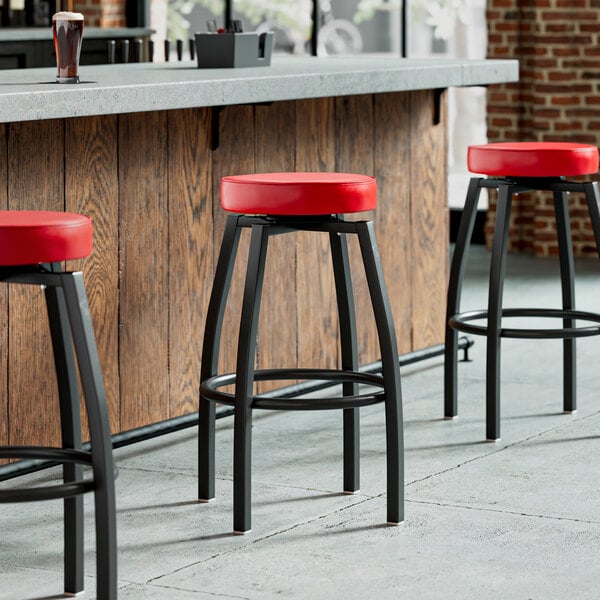 A row of Lancaster Table & Seating black bar stools with red seats in front of a bar.