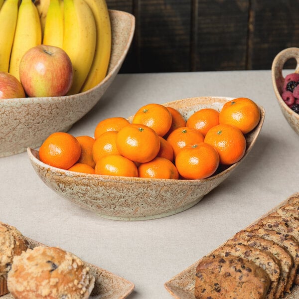 A 10 Strawberry Street Tiger Eye porcelain canoe bowl filled with oranges and bananas on a table with other bowls of fruit and cookies.