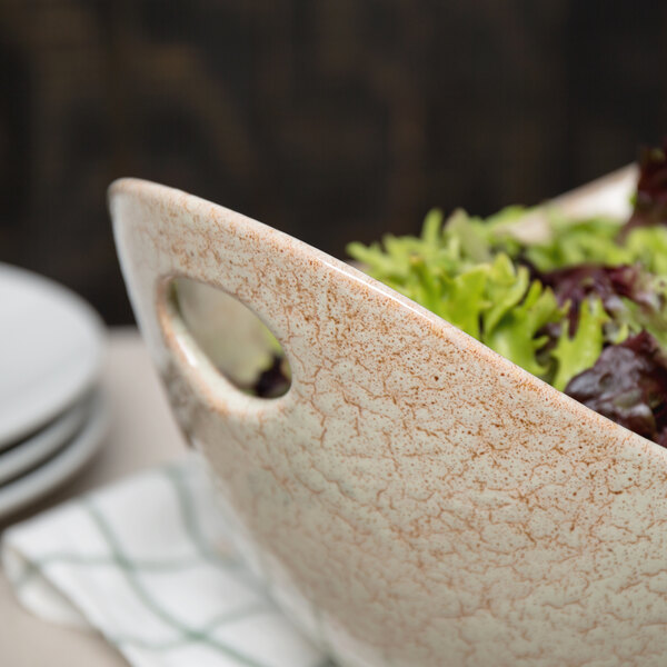 A white 10 Strawberry Street porcelain bowl with cut-out handles filled with lettuce and other food.
