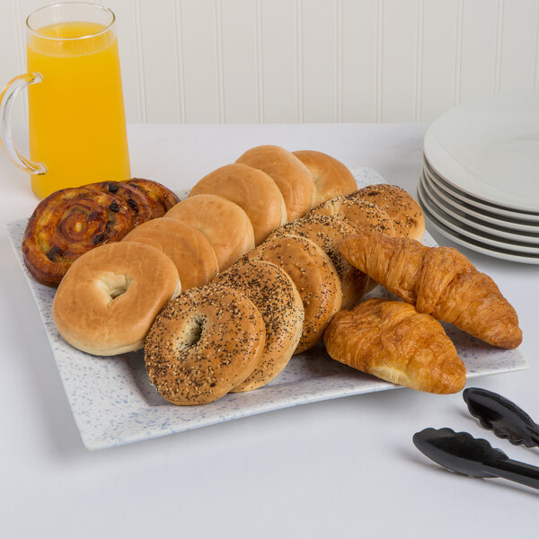 A 10 Strawberry Street blue speckled square porcelain platter with bagels, croissants, and orange juice on it.