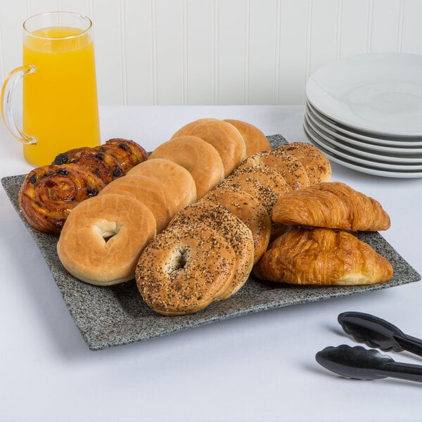 A 10 Strawberry Street granite square porcelain platter with bagels, croissants, and pastries on it, next to a glass of orange juice.