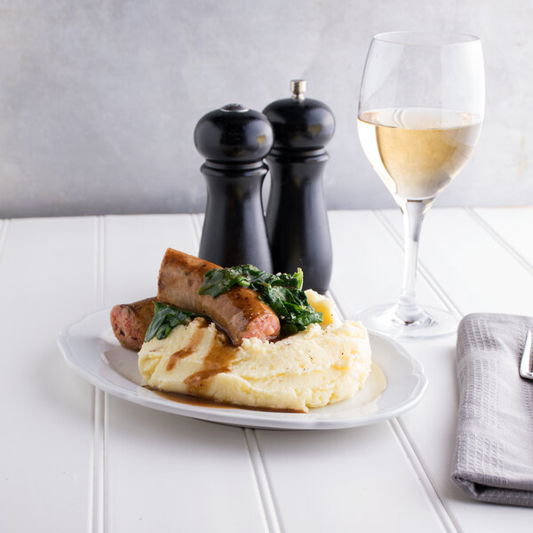 A Tuxton Charleston white china platter with sausage, mashed potatoes, and gravy on a table with a glass of white wine.