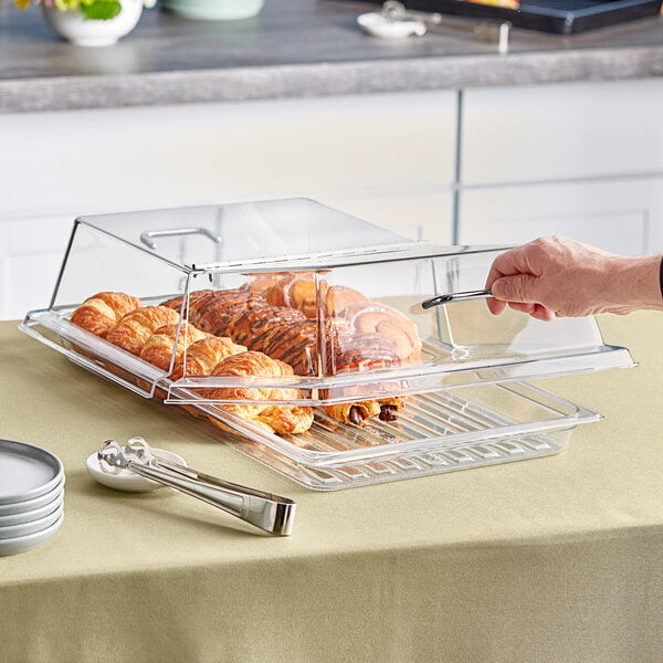 A person using a Cambro Clear Dome Display Cover with Hinged Lid to cover a tray of pastries on a counter.