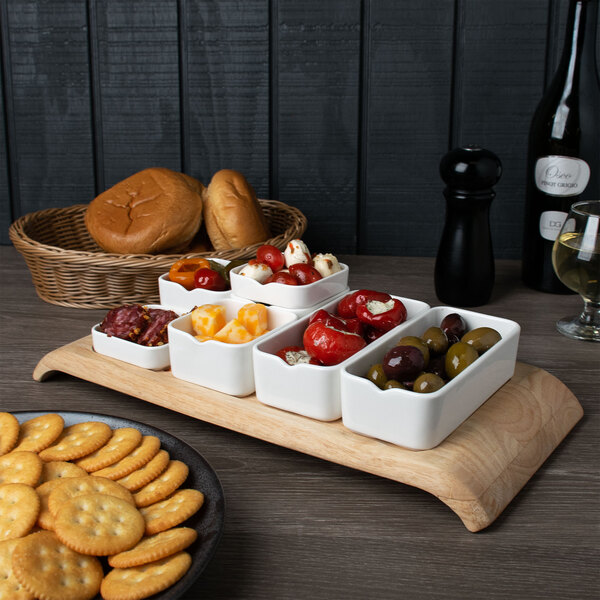 A wooden tray of food, including a white rectangular bowl filled with olives and peppers, on a table in a cocktail bar.