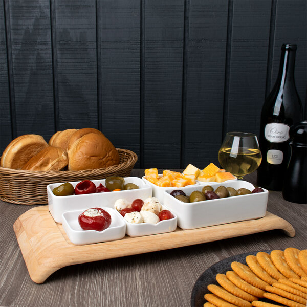 A white rectangular bowl filled with food on a table with a variety of food on a wooden tray.