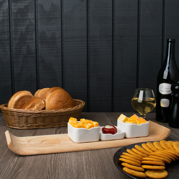 A white Arcoroc rectangular bowl filled with food on a wooden board with crackers and a glass of yellow liquid.