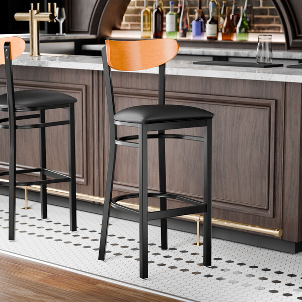 A black bar stool with a black vinyl seat and a cherry wood backrest.