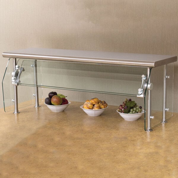 A stainless steel counter shelf with an Advance Tabco Sleek Shields food shield on it over bowls of fruit.