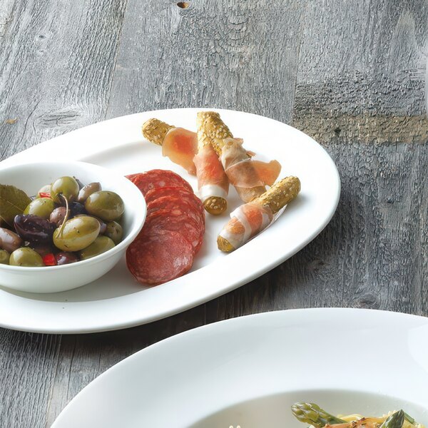 An oval white bone china platter with a plate of olives, salami, and vegetables on a table.