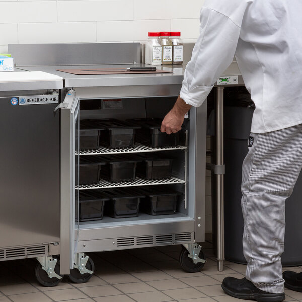 A man in a white shirt and gray pants opening a Beverage-Air worktop refrigerator in a school kitchen.