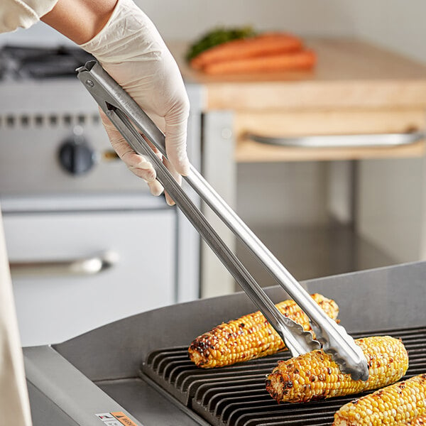 A person using Edlund heavy-duty tongs to cook corn on the cob on a grill.