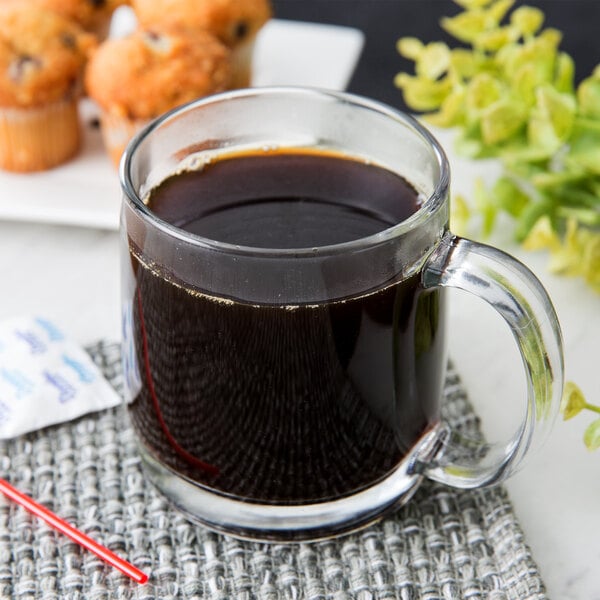 An Arcoroc tempered glass mug filled with brown liquid on a saucer with a cupcake.