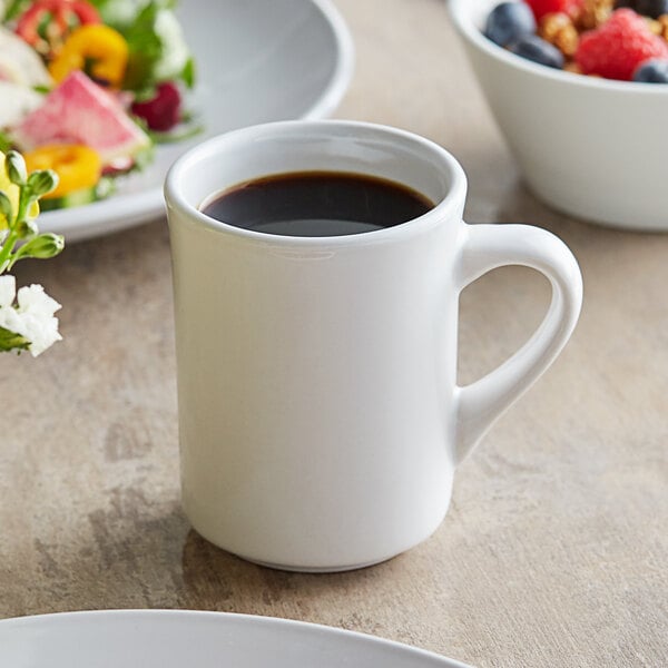 An Acopa ivory stoneware coffee mug filled with coffee on a table with a bowl of fruit.