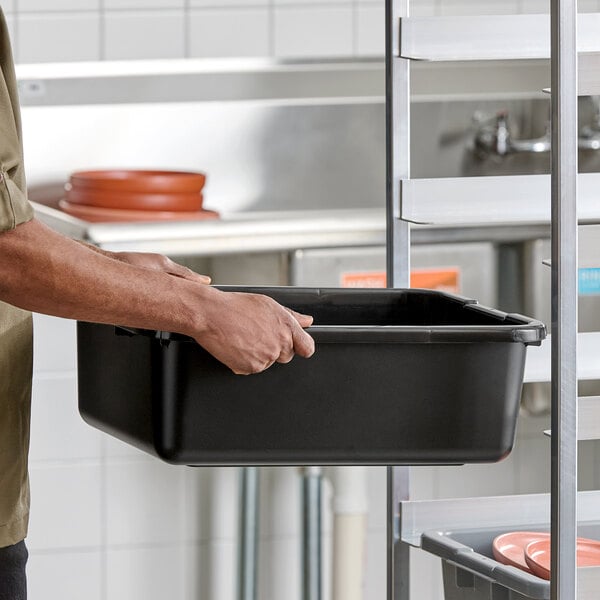 A black polyethylene bus tub with a ribbed bottom being held by a person in a commercial kitchen setting.