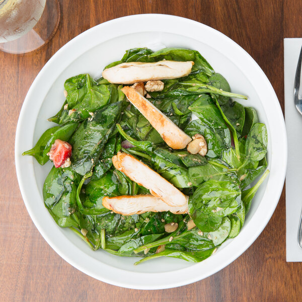 A white Arcoroc serving bowl filled with salad, chicken, and spinach on a table.