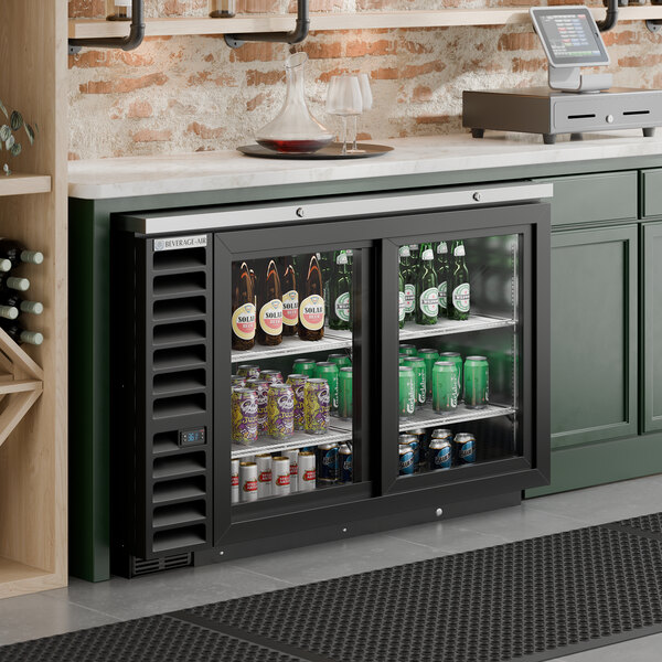 A black counter height sliding glass door back bar refrigerator filled with various bottled and canned beverages.