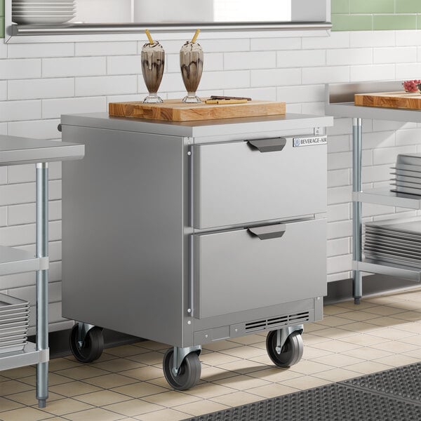 A compact stainless steel worktop refrigerator with two drawers and caster wheels.