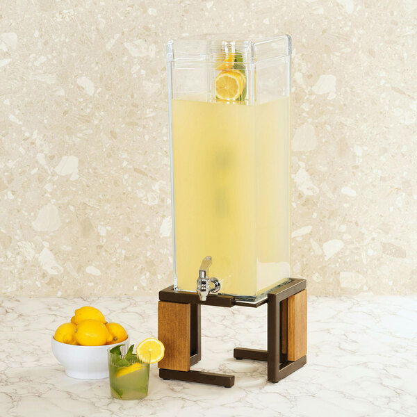 A Cal-Mil Sierra bronze and rustic pine beverage dispenser with a glass of yellow liquid and lemons.